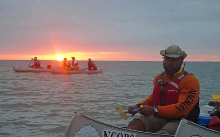 a group of people paddle canoes on open water as the sun sets on the horizon